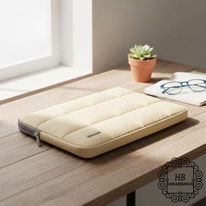 Può includere: Una custodia per laptop trapuntata color crema con una cerniera grigia e una piccola etichetta metallica. La custodia è rettangolare e si trova su una superficie di legno. Una piccola pianta grassa in vaso e degli occhiali sono sullo sfondo.