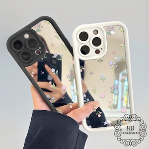 Puede incluir: Dos fundas para teléfono con diseños florales. Una funda tiene un marco negro y la otra un marco blanco. Ambas fundas tienen una superficie de espejo con pequeñas flores de colores. Las fundas se sostienen, mostrando el diseño y el reflejo.