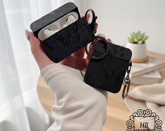 May include: Black AirPod case with a butterfly design. The case is open, revealing white AirPods inside. The case has a matte finish and a metal keyring attachment. The design includes embossed butterflies and text. The case is held in a person's hand.