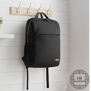 May include: A black backpack with a minimalist design hangs on a wooden rack with metal hooks. The backpack has a rectangular shape, a front pocket, and a top handle. The zipper is gold-colored, and the bag has two black straps.