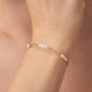Puede incluir: Un delicado brazalete de cadena dorada con pequeñas perlas blancas. El brazalete está en la muñeca de una persona.