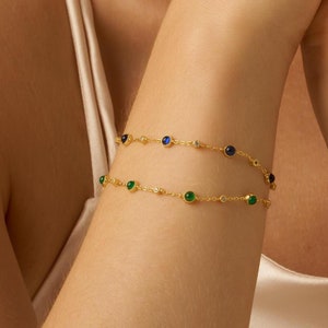 May include: Two gold chain bracelets with small green and blue gemstones. The bracelets are layered on a person's wrist.