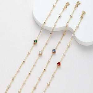 May include: Gold chain bracelets with coloured square gemstones and small pearl accents. Each bracelet has a different gemstone colour: green, clear, blue, and red. The bracelets are displayed on a white surface.