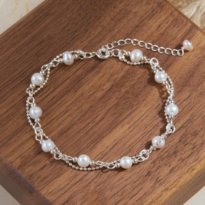 以下が含まれることがあります： 小さな真珠が2連になった、繊細なシルバーブレスレット。ブレスレットは留め金で留められており、木製の表面に置かれています。