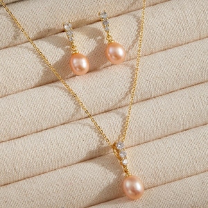 以下が含まれることがあります： ゴールドカラーのネックレスとイヤリングのセットで、ピーチカラーのパールが特徴です。ネックレスにはシングルパールのペンダントがあり、イヤリングにはそれぞれパールのドロップが付いています。ジュエリーはベージュのジュエリーボックスに展示されています。