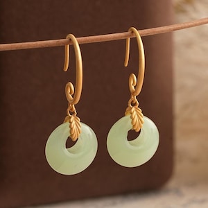 May include: A pair of gold-toned earrings featuring a matte finish hook with a spiral detail at the top. Each earring suspends a smooth, pale green circular stone pendant attached by a small, textured gold-toned leaf-shaped metal connector.
