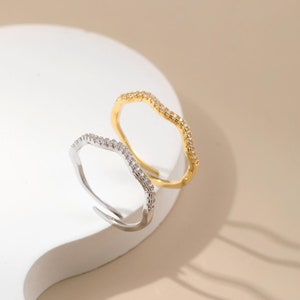 Könnte beinhalten: Zwei wellenförmige Ringe, einer silberfarben und einer goldfarben, werden auf einer weißen Oberfläche präsentiert. Der silberne Ring hat eine Reihe kleiner, klarer Steine. Der goldfarbene Ring hat ein ähnliches Design mit klaren Steinen.