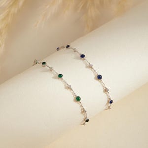 Può includere: Un delicato bracciale a catena in argento con piccole pietre rotonde alternate verdi e blu incastonate in metallo, intervallate da minuscole pietre trasparenti lungo la catena, creando un gioiello raffinato e colorato.