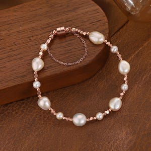 May include: A rose gold bracelet featuring a series of white, irregularly shaped pearls interspersed with small, round beads and square-cut accents. The bracelet has a magnetic clasp and a delicate chain for added security. The pearls vary in size, creating a unique and elegant design.