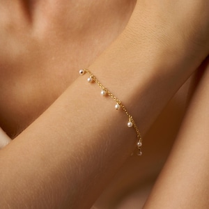 May include: A delicate gold chain bracelet with small white pearls dangling from it. The bracelet is shown on a person's wrist.