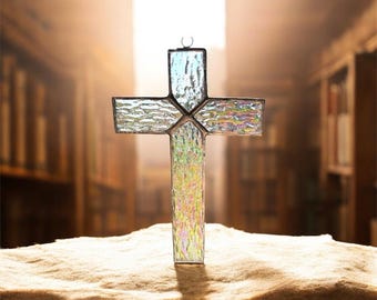 May include: A stained glass cross featuring a textured, iridescent surface with a metallic border. The cross has a small circular loop at the top for hanging and displays a translucent, multi-colored finish that catches light across its panels.
