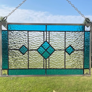 May include: A rectangular stained glass panel featuring a geometric design with clear textured glass panes framed by vibrant teal and blue borders. The piece includes three central diamond-shaped accents and is suspended by a silver metal chain.