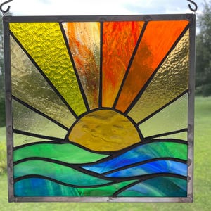 May include: A square stained glass panel featuring a sunrise over water. The sun is a textured yellow semicircle with radiating beams in yellow, orange, and red. Below, the water is depicted with wavy green and blue glass segments.