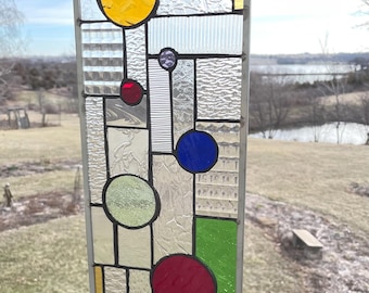 May include: A rectangular stained glass panel with a geometric design. It features various shapes, including circles and rectangles, in colors like yellow, red, blue, and green. The glass has textured and clear sections, framed with a metal border. The panel is hanging outdoors.