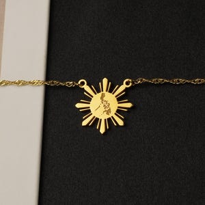 May include: A gold-toned necklace featuring a sunburst-shaped pendant with a central cutout map silhouette. The pendant is attached to a delicate twisted chain, creating a metallic accessory that rests horizontally across the frame.