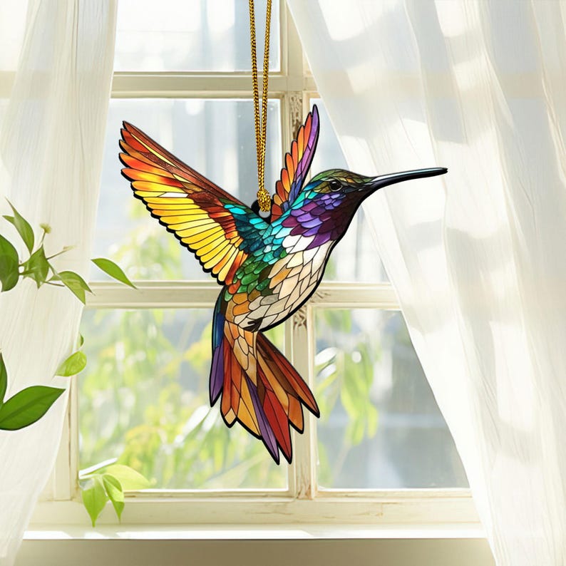 May include: A stained glass hummingbird ornament with a gold cord hanging from a window. The hummingbird is multi-colored with shades of yellow, orange, red, green, blue, and purple.