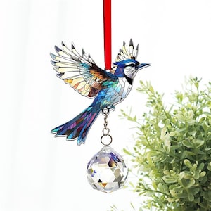 May include: A stained glass blue jay ornament with a crystal ball hanging from it. The ornament is suspended from a red ribbon.