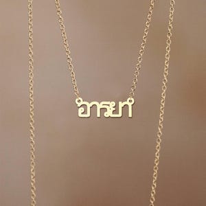 May include: A gold-coloured necklace featuring the Thai word "อารยา" as a pendant. The pendant is in a sans-serif font. The delicate chain hangs against a neutral background.