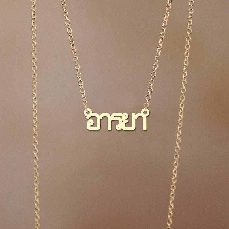 May include: A gold-coloured necklace featuring the Thai word "อารยา" as a pendant. The pendant is in a sans-serif font. The delicate chain hangs against a neutral background.