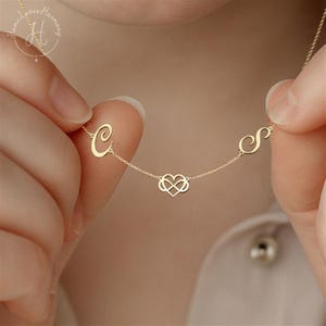 May include: A gold-toned necklace featuring a delicate chain with a central infinity heart charm flanked by two cursive script letters, C and S. The metal components are polished and connected by thin links, creating a minimalist jewelry piece.