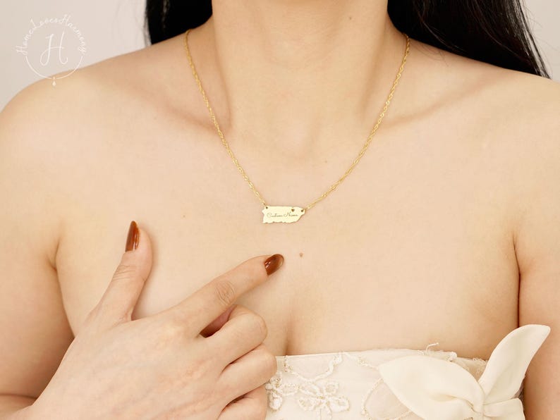 May include: A gold-toned necklace featuring a small, flat pendant shaped like the island of Puerto Rico. The pendant is engraved with cursive text and a tiny heart symbol. The necklace is attached to a delicate, thin gold-toned chain.