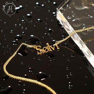 May include: A gold-toned metal necklace featuring a nameplate pendant with the script text Sølvi. The delicate chain is attached to both sides of the nameplate, which is displayed against a dark, reflective surface with scattered water droplets.