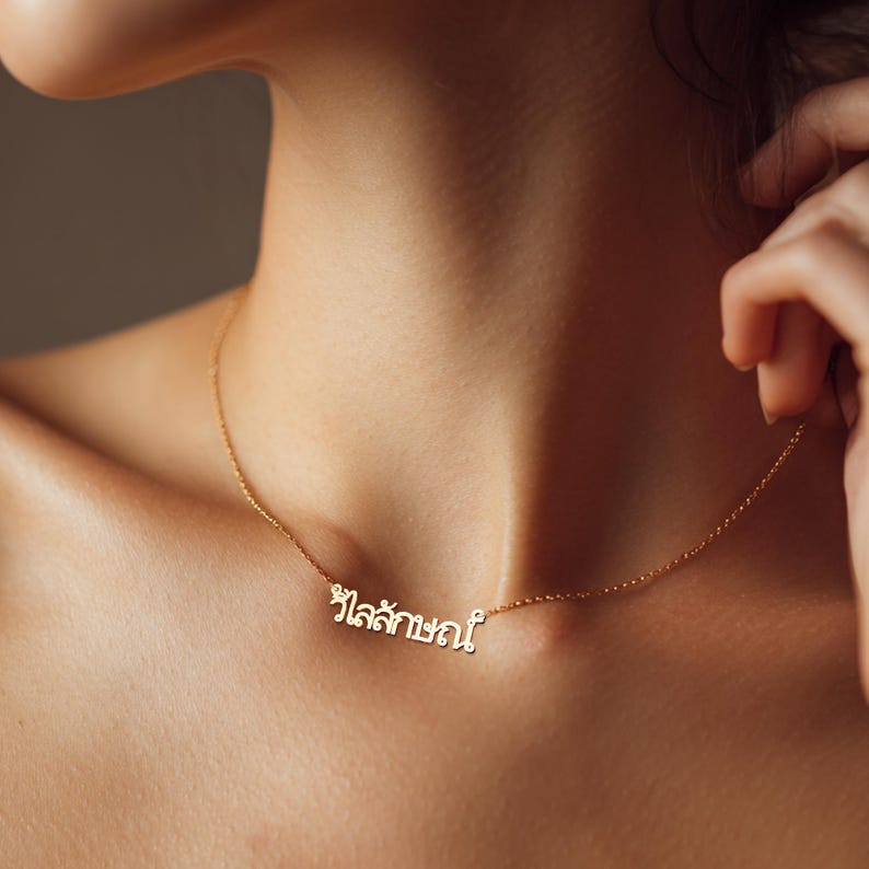 May include: A delicate gold necklace featuring the Thai word "วิไลลักษณ์" as a pendant. The necklace is worn around the neck. The gold chain is thin and the pendant is in a cursive font. The chain rests on the skin.