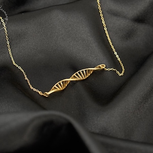 May include: A gold-toned necklace featuring a central pendant shaped like a double helix DNA strand. The pendant is connected to a delicate gold-toned chain. A circular logo with the text "Homes Loves Harmony" is visible in the upper left corner.