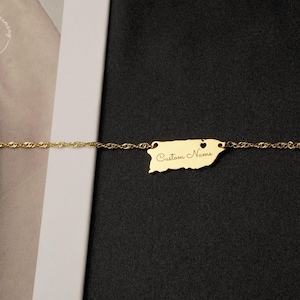 May include: A gold-colored chain necklace featuring a pendant shaped like the island of Puerto Rico. The pendant includes a small heart cutout and the engraved text Custom Name in a cursive script font, centered on the metallic surface.