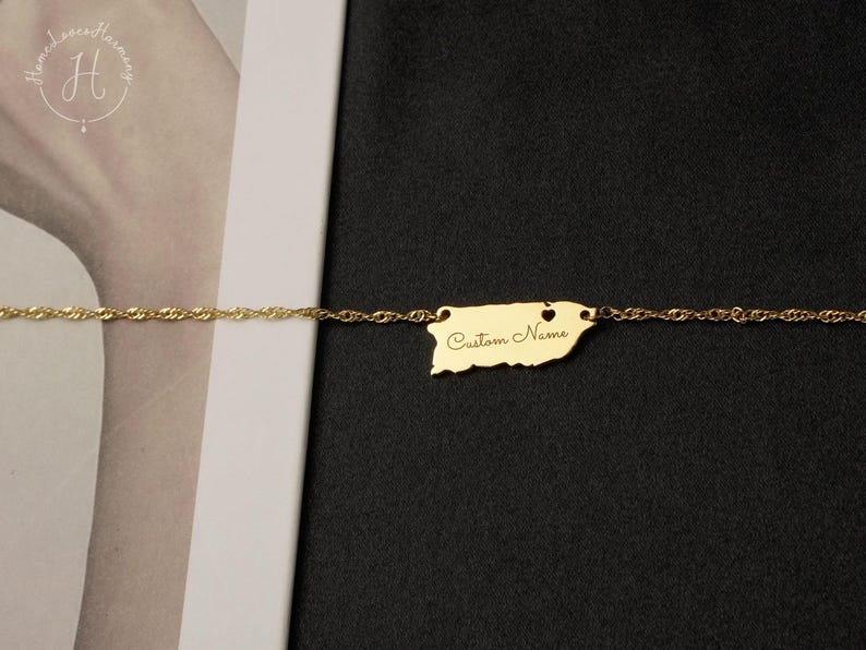 May include: A gold-colored chain necklace featuring a pendant shaped like the island of Puerto Rico. The pendant includes a small heart cutout and the engraved text Custom Name in a cursive script font, centered on the metallic surface.