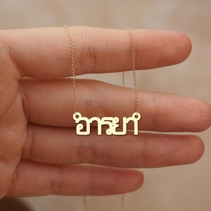 May include: A gold-coloured necklace with a pendant displaying Thai script. The pendant hangs from a fine chain. The necklace is held up by a hand, highlighting the jewellery's design and colour.