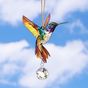 May include: A stained glass hummingbird with iridescent colors hangs from a string with a crystal prism hanging below. The hummingbird is in flight with its wings spread out.