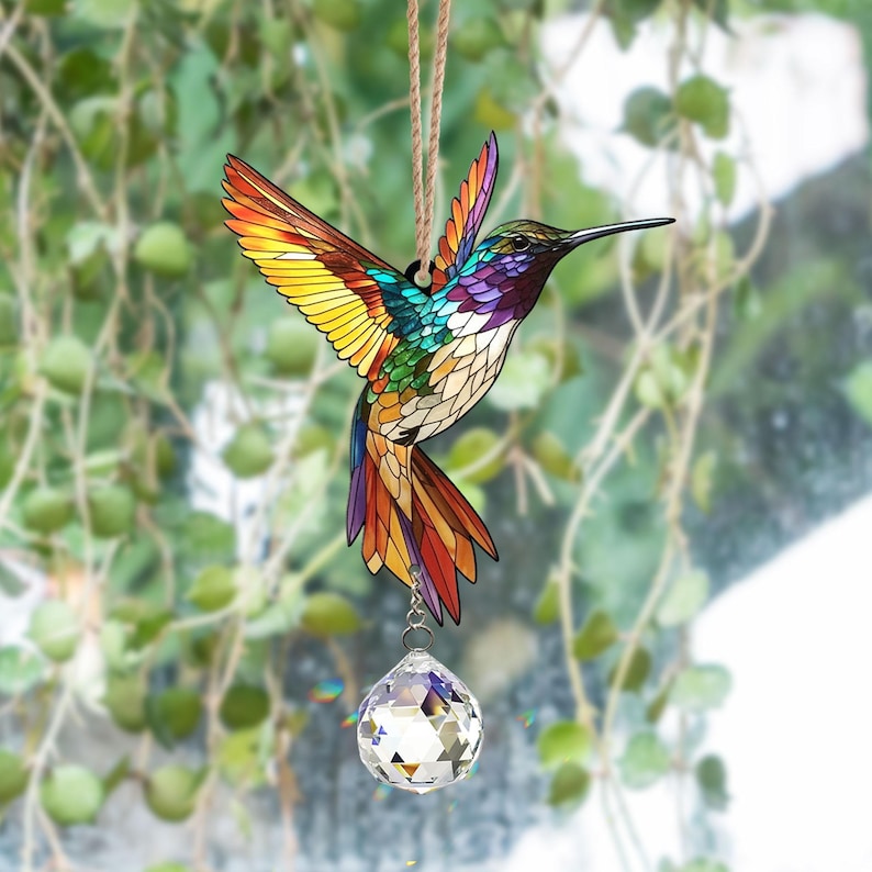 May include: A stained glass hummingbird suncatcher with a crystal prism hanging from it. The hummingbird is colorful with shades of yellow, orange, green, blue, and purple. The crystal prism is clear and reflects light.