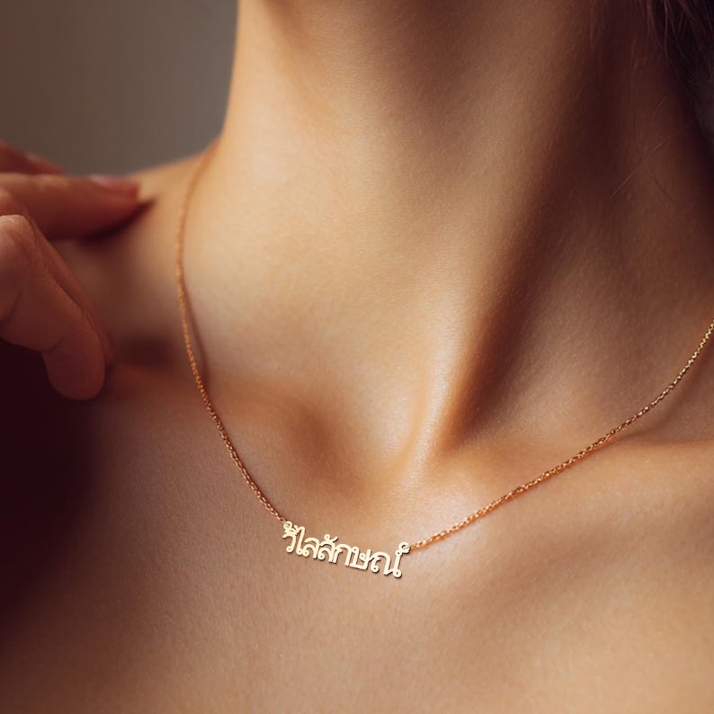 May include: A delicate gold necklace featuring a Thai script pendant. The necklace is worn around the neck, with the pendant resting on the chest. The chain is fine and the pendant is small, with the Thai script in a simple font.