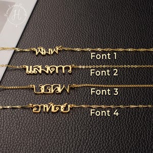 May include: Four gold-colored necklaces with custom Thai script nameplates, each featuring a distinct chain style and font labeled Font 1, Font 2, Font 3, and Font 4. The necklaces are displayed horizontally against a black textured background.