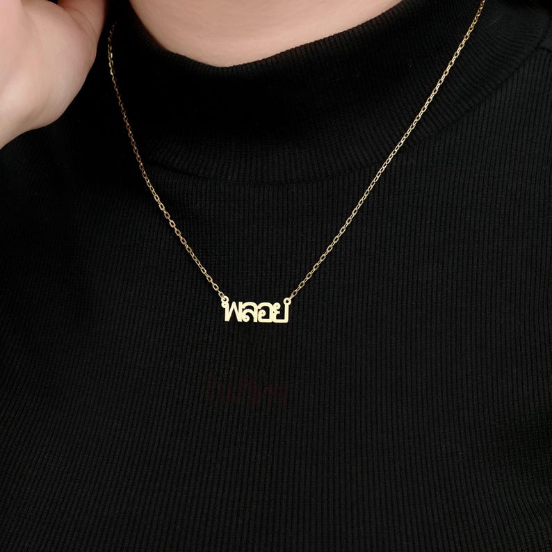 May include: A delicate gold-coloured necklace with a chain and a pendant spelling out a word in Thai script. The necklace is displayed against a black background.