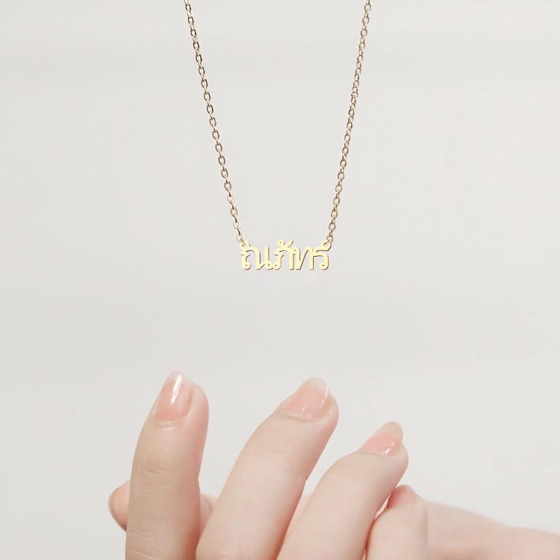 May include: A delicate gold necklace with a chain and a pendant featuring Thai script. The necklace is displayed against a soft, neutral background. The chain is fine and the pendant is small, creating an elegant and minimalist aesthetic.