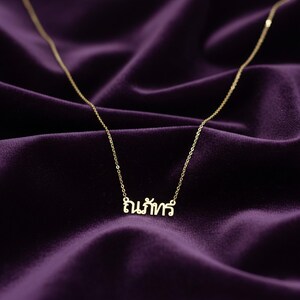 May include: A delicate gold necklace with a chain and a pendant spelling out a word in a script font. The necklace is displayed on a deep purple velvet surface.