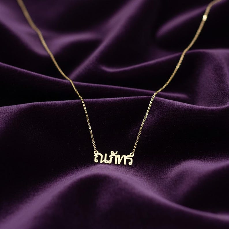May include: A delicate gold necklace with a chain and a pendant spelling out a word in a script font. The necklace is displayed on a deep purple velvet surface.