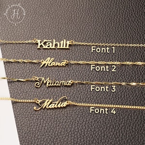 May include: Four gold-toned name necklaces displayed horizontally, each featuring a distinct script or block font style labeled Font 1, Font 2, Font 3, and Font 4. The necklaces showcase different chain link designs, including cable, twisted, and flat bar styles.