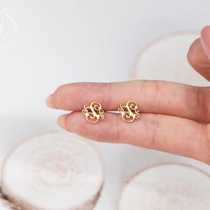 May include: A pair of gold-toned stud earrings featuring an intricate, openwork monogram design with swirling, decorative flourishes. The earrings are crafted with a polished finish and display a detailed, ornate letter shape in a metallic gold color.