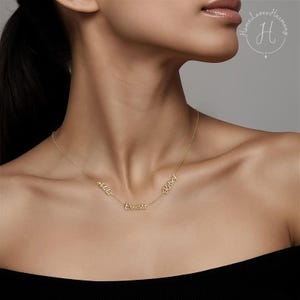 May include: A gold-toned necklace featuring three distinct name pendants in a cursive script font. The names Adela, Ernesta, and Kelsey are integrated into the delicate chain, creating a layered look across the collarbone area of the neck.