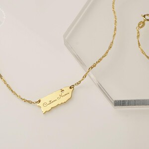 May include: A gold-toned necklace featuring a delicate twisted chain and a flat, map-shaped pendant. The pendant is engraved with the text "Custom Name" in a cursive script and includes a small heart cutout near the top edge.