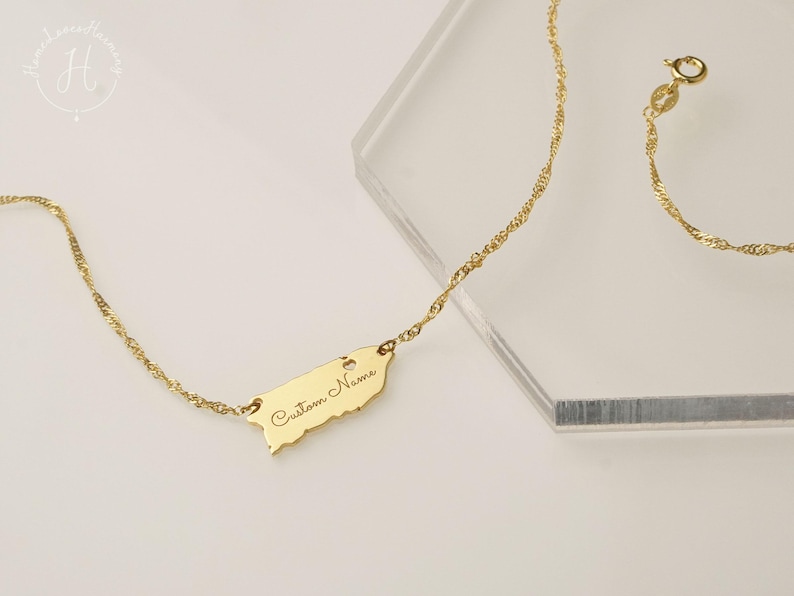 May include: A gold-toned necklace featuring a delicate twisted chain and a flat, map-shaped pendant. The pendant is engraved with the text "Custom Name" in a cursive script and includes a small heart cutout near the top edge.