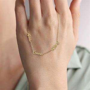 May include: A gold-toned necklace featuring three distinct names, Adela, Ernesta, and Kelsey, written in a cursive script font. The names are integrated directly into the delicate chain, creating a personalized piece of jewelry with a polished finish.