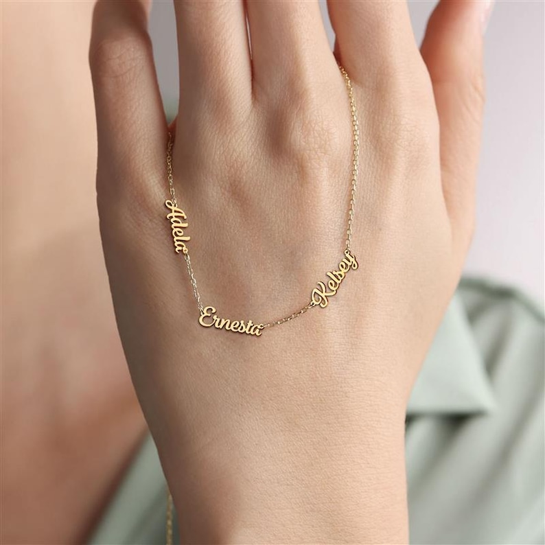 May include: A gold-toned necklace featuring three distinct names, Adela, Ernesta, and Kelsey, written in a cursive script font. The names are integrated directly into the delicate chain, creating a personalized piece of jewelry with a polished finish.