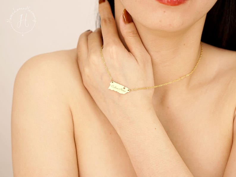 May include: A gold-toned necklace featuring a delicate chain and a small, flat pendant shaped like the island of Puerto Rico. The pendant is engraved with cursive text and a tiny heart symbol, suspended from a thin, textured link chain.