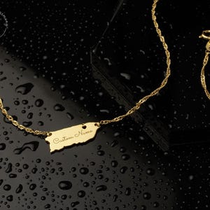 May include: A gold-colored necklace featuring a pendant shaped like the island of Puerto Rico. The pendant includes a small heart cutout and the engraved text Custom Name. The chain is a twisted link style with a lobster claw clasp.