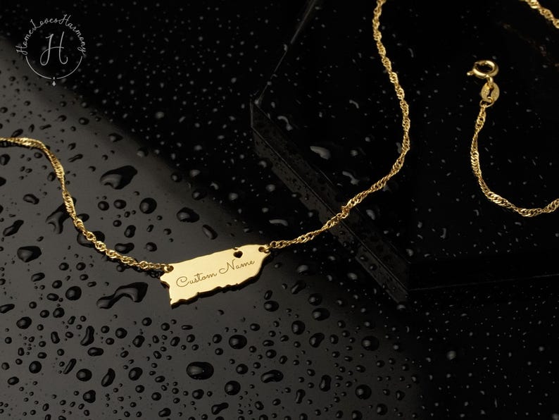May include: A gold-colored necklace featuring a pendant shaped like the island of Puerto Rico. The pendant includes a small heart cutout and the engraved text Custom Name. The chain is a twisted link style with a lobster claw clasp.