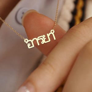 May include: A delicate gold necklace with a pendant bearing Thai script. The pendant hangs from a fine chain. The necklace is held by a person.